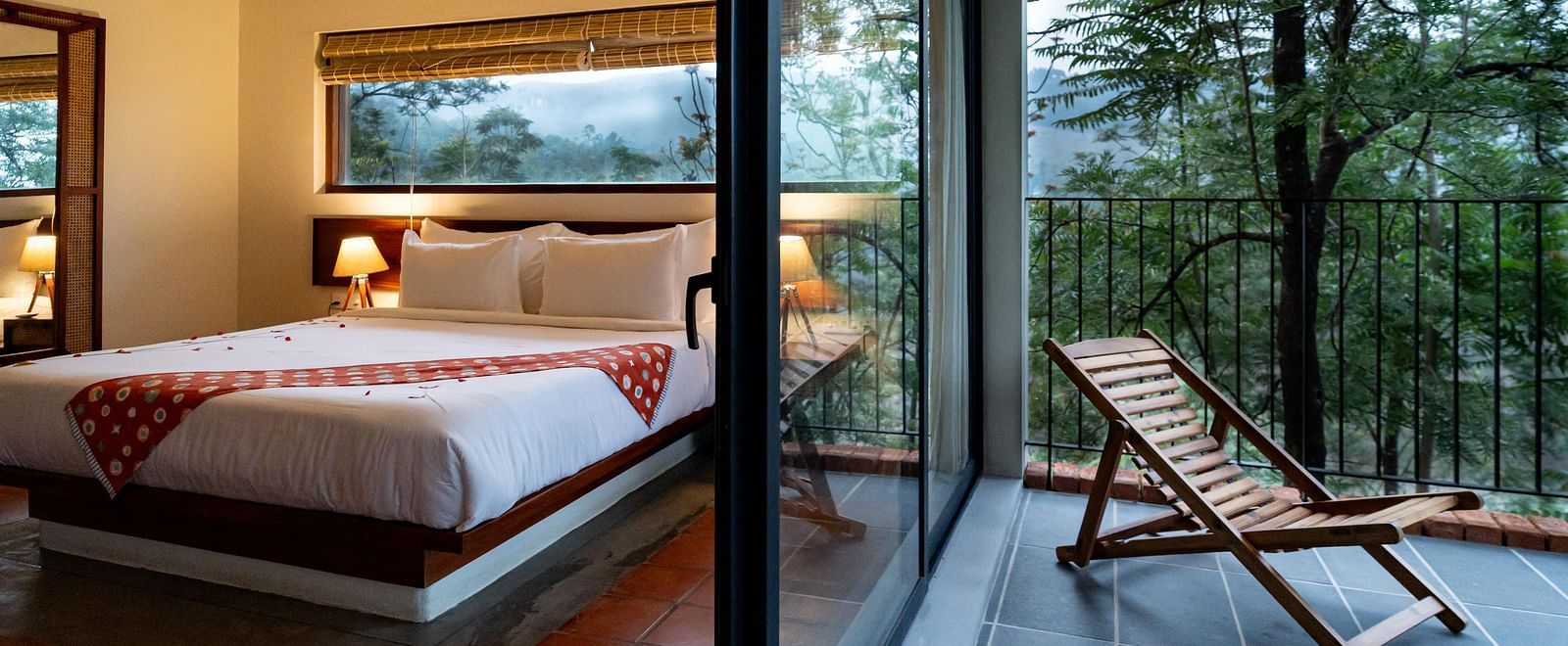 A spacious room at Vaasta Vagamon with a comfortable bed, leading to a private balcony with a wooden lounge chair and scenic views.