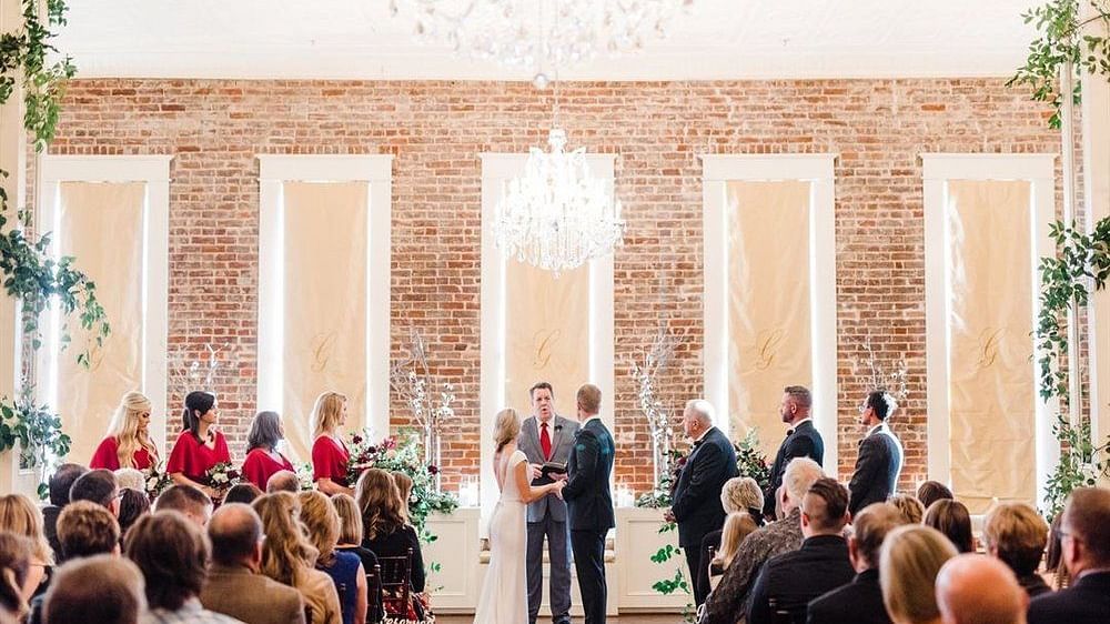 A grand wedding ceremony in a luxurious hall with tall windows, chandeliers, and an audience watching the couple exchange vows.- The Grand Hotel & Ballroom, McKinney