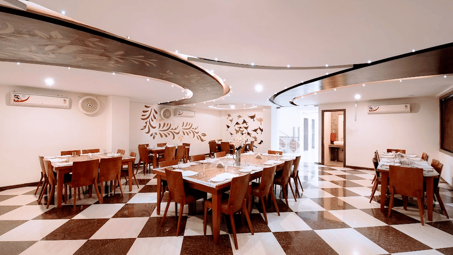 Spacious dining hall with dark wood tables, chairs, curved false ceiling details, and a checkered tiled floor.