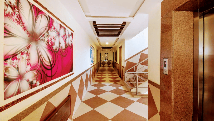 Well-lit hotel corridor next to an elevator, featuring patterned wall tiles and a large pink floral painting.