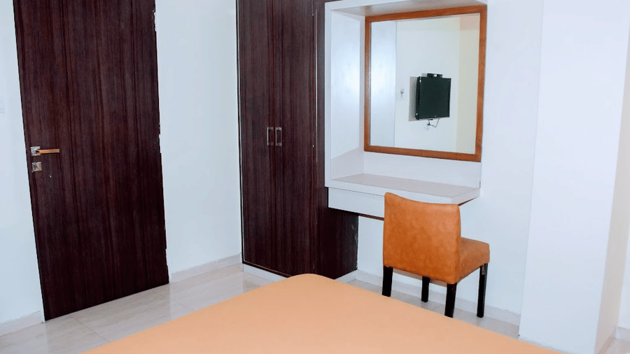 Deluxe room interior featuring a wooden door, dark brown wardrobe, vanity mirror desk, and an orange chair.