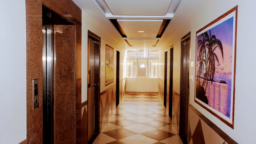 A well-lit hotel elevator hallway with a tiled diamond-pattern floor, wooden wall panels, and framed art.