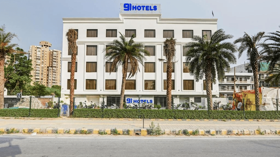 Facade of Hotel 91 Privé, Gurugram, with equidistant palm trees on its perimeter against a clear blue sky in the background.