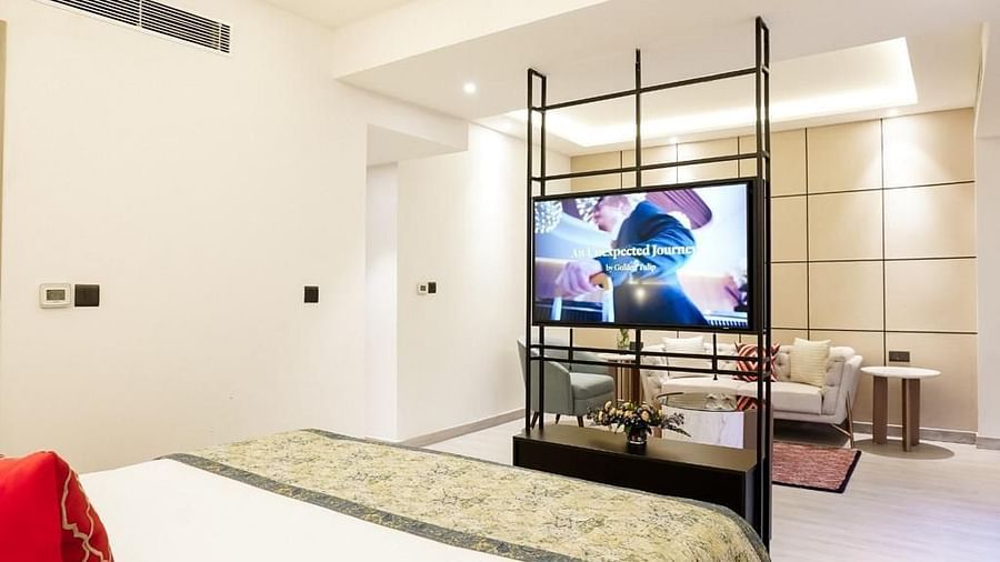 Modern hotel suite with a comfortable bed featuring patterned bedding and red cushions, a TV mounted on a sleek open-frame partition, and a cozy seating area with a beige sofa and accent pillows in the background - Golden Tulip, Lucknow