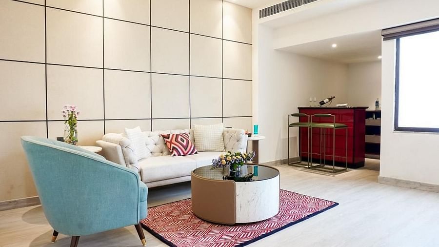 A living area with a beige sofa adorned with decorative cushions, a teal accent chair, a modern round coffee table with fresh flowers, and a compact bar counter with high stools in the background - Golden Tulip, Lucknow