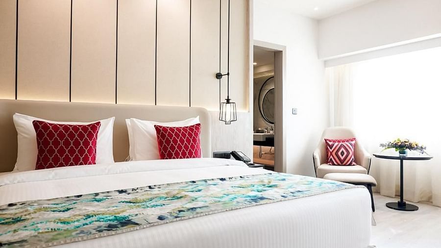 Bright and modern hotel room with a king size bed, red patterned cushions, pendant lighting, and a cosy window-side seating area with a table and flower arrangement - Golden Tulip, Lucknow