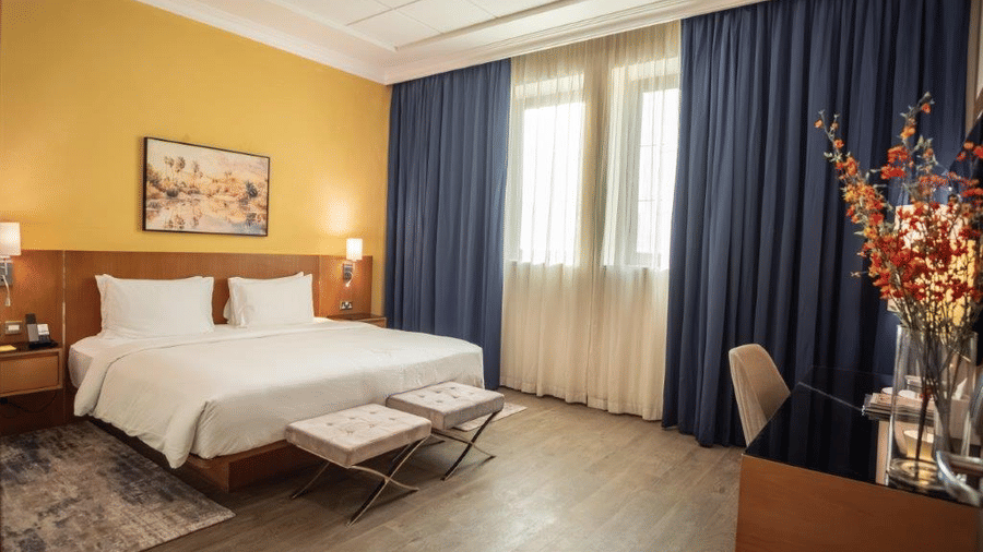 Well-lit bedroom with a king-size bed, desk, chair, and decorative elements at La Maison Hotel, Doha.
