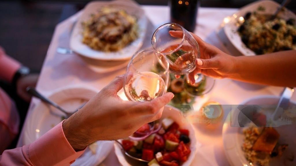 A close-up of a romantic dinner setting with two pairs of hands clinking wine glasses over a table with plates of food, candles, and a soft, warm glow.