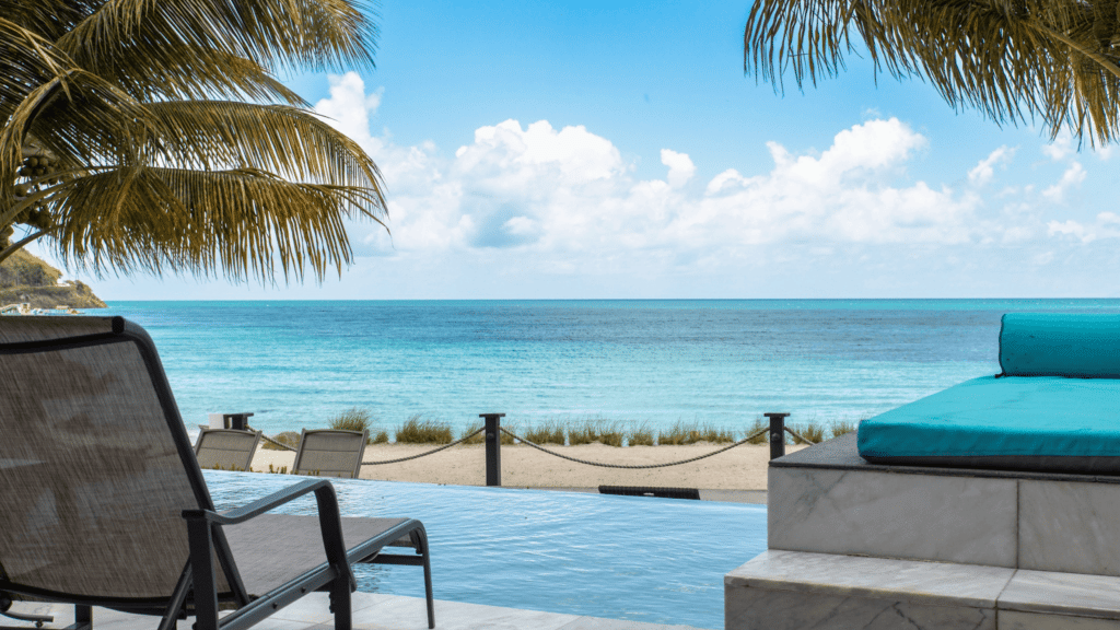 An outdoor patio area with lounge chairs overlooks a pristine turquoise beach and ocean under a clear sky.
