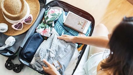 A person packing a suitcase with clothing, accessories like a sun hat, sunglasses, and a wallet, preparing for a trip.