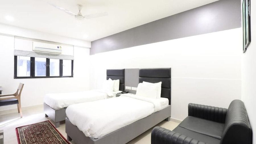 Corner view of the Premium Tin Room at Pravasa Stay, Ahmedabad (Alba Premier), showcasing twin beds along wit a patterned rug in front of it and a black leather couch kept beside it.