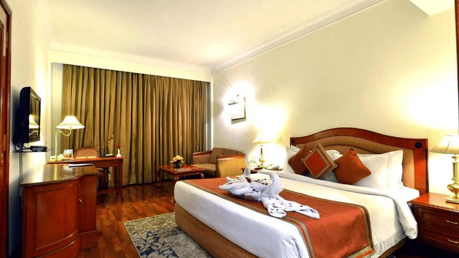 Standard Room at The Bristol Hotel, Gurgaon with a king-size bed, rust-coloured runner, a towel swan decoration, a wooden desk, an armchair, and curtains.