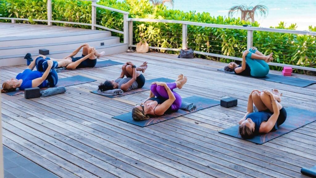 Several people are engaged in a yoga or fitness class on a wooden deck overlooking the ocean.