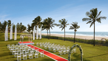 Outdoor beach wedding setup on a lawn, with white chairs, a red aisle, floral arch, palm trees, and the Arabian Sea in the background - Caravela Beach Resort, Goa