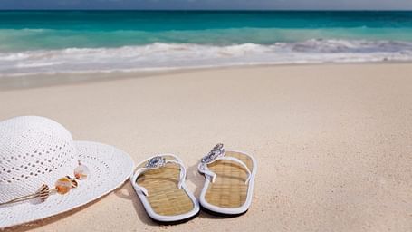 Image showing a white hat, slippers on the sand with ocean waves in the background.