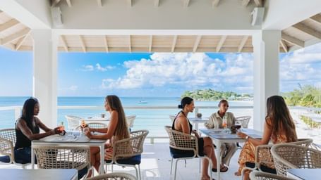 Guests dining at an open-air restaurant with white pillars and a high ceiling, enjoying a panoramic view of the sparkling blue ocean and distant land under a partly cloudy sky.