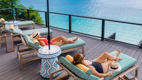 Two women lounging on sunbeds under a yellow umbrella on a wooden deck at Tamarind Hills Resort, Antigua, overlooking the turquoise Caribbean sea.