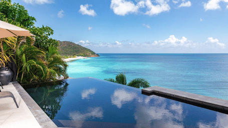 An infinity pool at the Tamarind Hills Resort and Villas, Antigua, with a lounge chair by the poolside, and ocean view in the front and green mountains in the background.