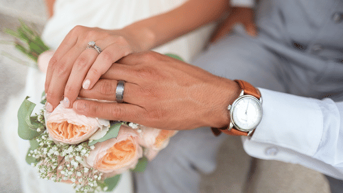 A close-up of a bride and groom's hands, displaying wedding rings and a bouquet of pale roses.
