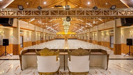 A large banquet hall with a stage area, speakers, and seating at the rear of Angel Lords Eco Inn Amreli.