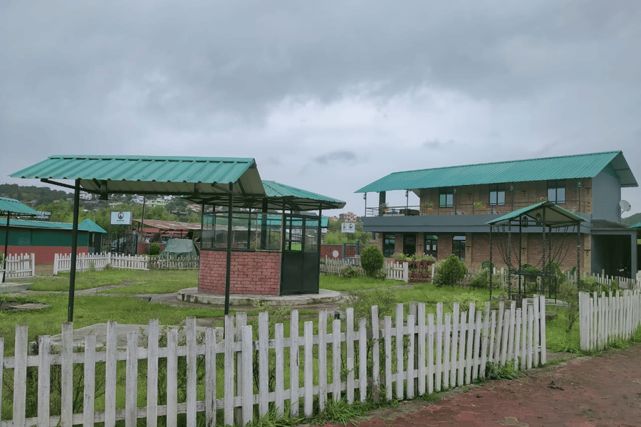 A facade of Woodstock Farmhouse, Shillong, featuring buildings, fence, and sky in the backdrop.