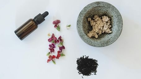A glass vial, flowers, herbs and a mortar aesthetically placed on a white surface.