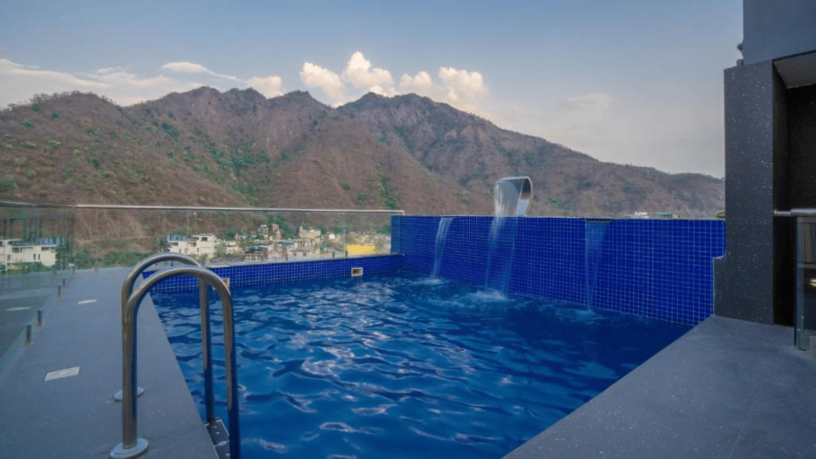 An outdoor swimming pool with metal handrails, water flowing from a spout, and a mountain range visible in the background under a clear sky at Perfectstayz Premium Laxmi Heritage.
