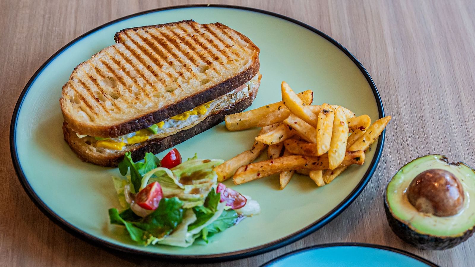 One plate featuring a sandwich with veggies on the side and crispy French fries, while another plate holds an omelette with a half avocado placed beside both plates at Indie Stays Mumbai International Airport.