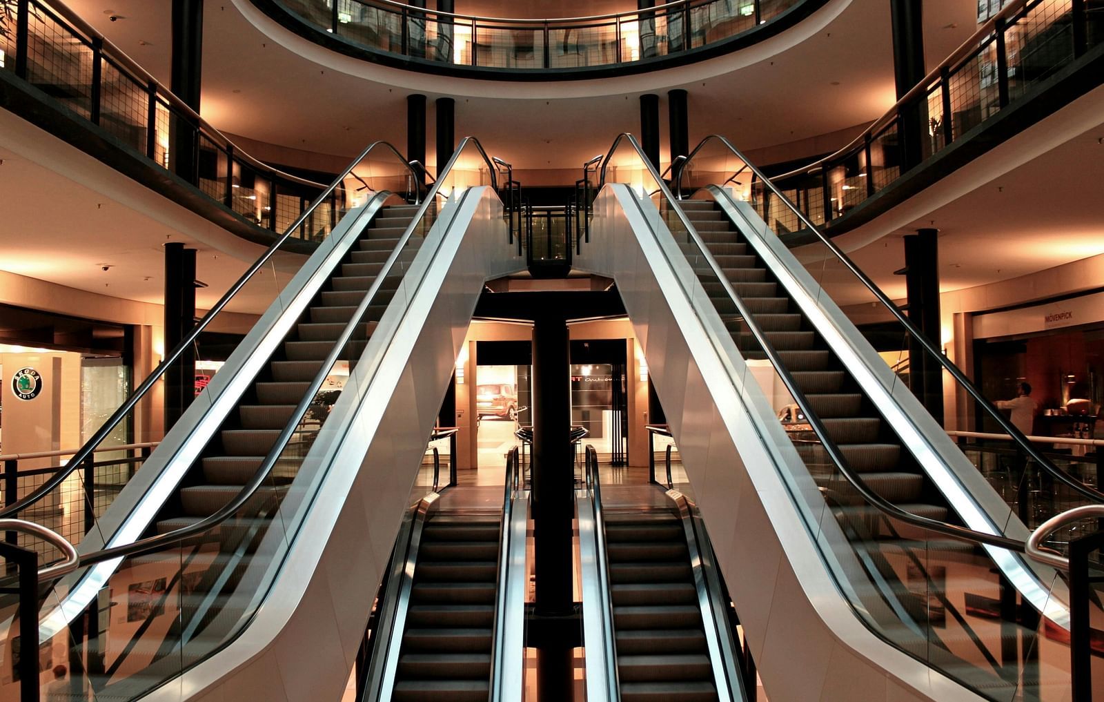Interior view of a shopping mall with two escalators ascending side-by-side, surrounded by shops on multiple levels and warm lighting from spherical lights.
