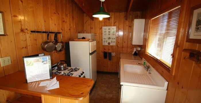 A compact cabin kitchen with warm wood paneling featuring a small sink, a wooden countertop with a menu and utensils. - Kit Carson Lodge