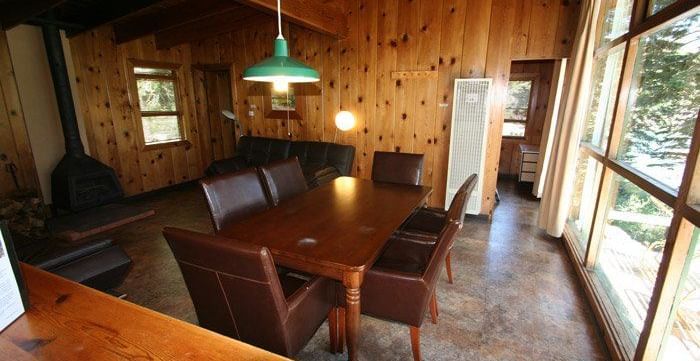 A rustic cabin interior with wood paneling featuring a dining table and hairs. - Kit Carson Lodge