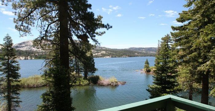 A scenic lake view framed by pine trees and a green railing, with distant mountains under a blue sky.- Kit Carson Lodge & Restaurant 