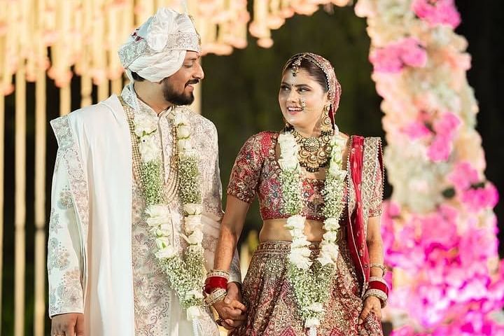 A newlywed couple walks hand in hand, exchanging joyful smiles, adorned in traditional attire with floral garlands. - Umaid Palace - A Lakeside Heritage Palace Resort