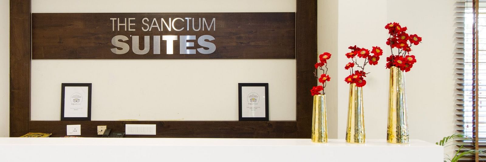 A reception area is showing 'The Sanctum Suites' sign and including decorative red flowers in vases placed on the desk at Sanctum Suites Whitefield, Bangalore.