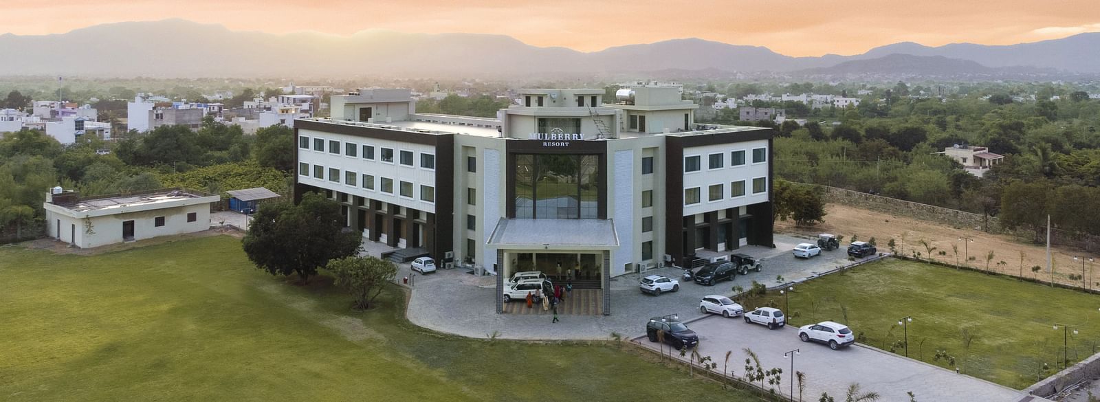 An aerial view of the Bamboo Saa Mulberry Resort, Pushkar at sunset or sunrise surrounded by green lawn and the backdrop of hills.