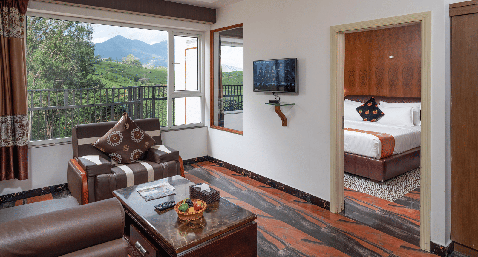 The spacious interior of the Premium Room at Parakkat Nature Hotel and Resorts, Munnar featuring a separate living area with wooden furnishing, an opening towards bedroom, and large glass windows overlooking the greenery.