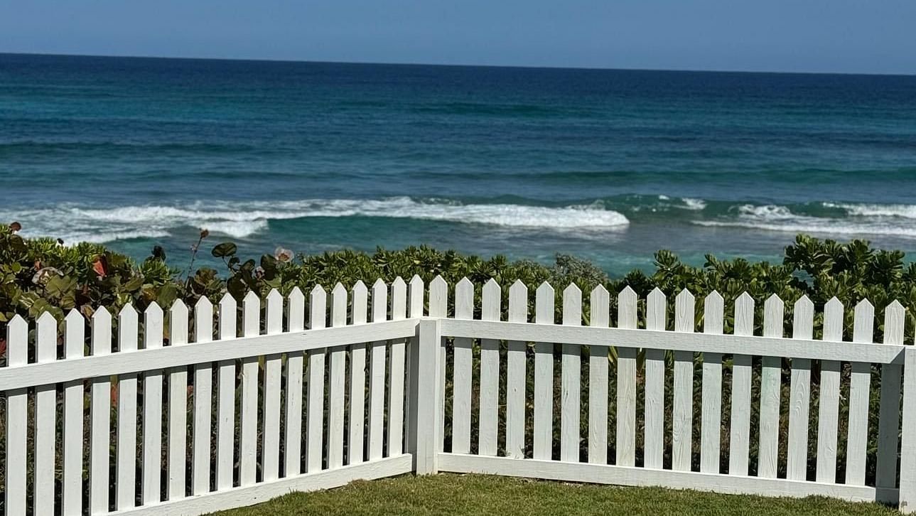 The ocean with small waves is visible beyond a well-maintained lawn enclosed by a picket fence. The sky is a clear | Abaco Inn