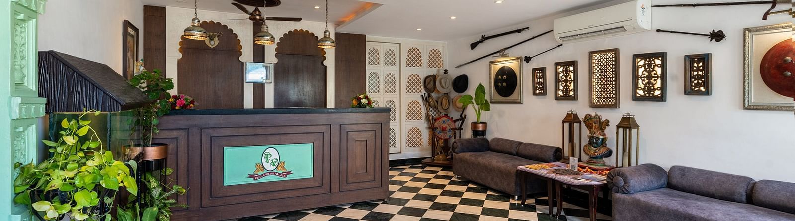 Reception area at Panna Vilas Palace features a wooden reception desk, walls adorned with wall-mounted frames, and several potted plants | Panna Vilas Udaipur