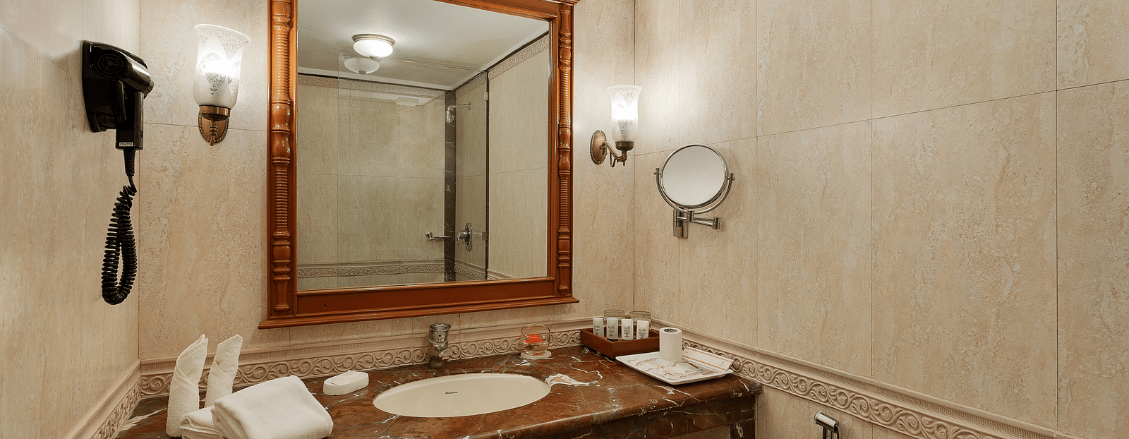 A spacious bathroom with a large mirror, a marble vanity, and a toilet. The walls are decorated with wallpaper and there is a wall-mounted phone. 