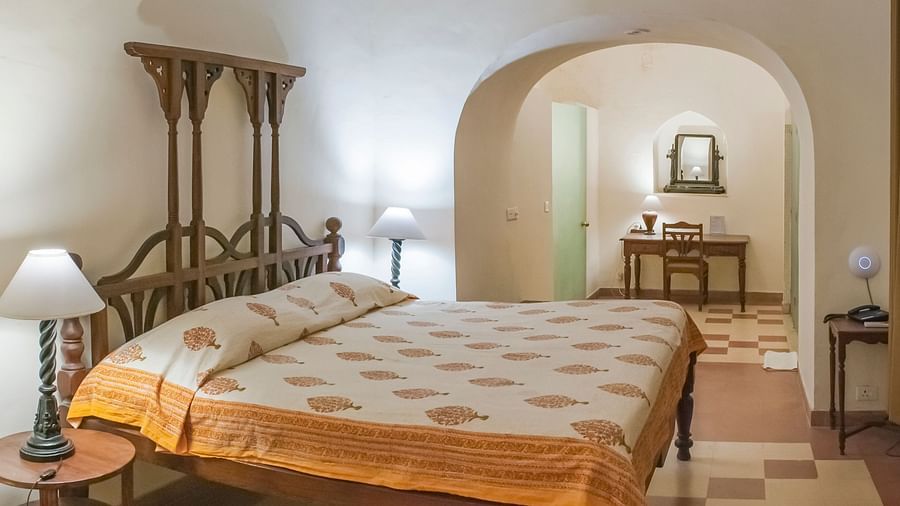 Bedroom with a bed, decorative headboard, bedside tables with lamps, and an arched opening to another room with a desk at Tijara Fort-Palace, Alwar.