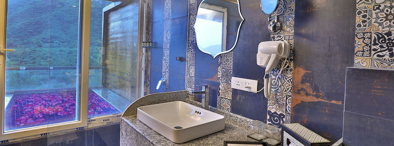 A bathroom with a unique patterned tile design, a sink on a speckled counter, and a window overlooking a lush landscape.- Bamboo Saa Resort & Spa, Udaipur