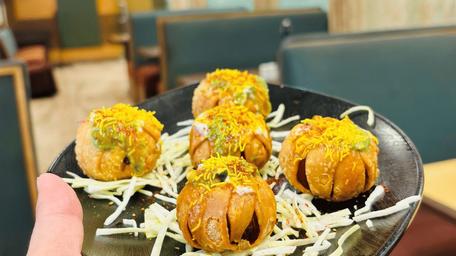 A person holding a plate with 5 pieces of an Indian fritters dish, with the seating area of the Navidya Restaurant  as the backdrop - Amantra Comfort Hotel