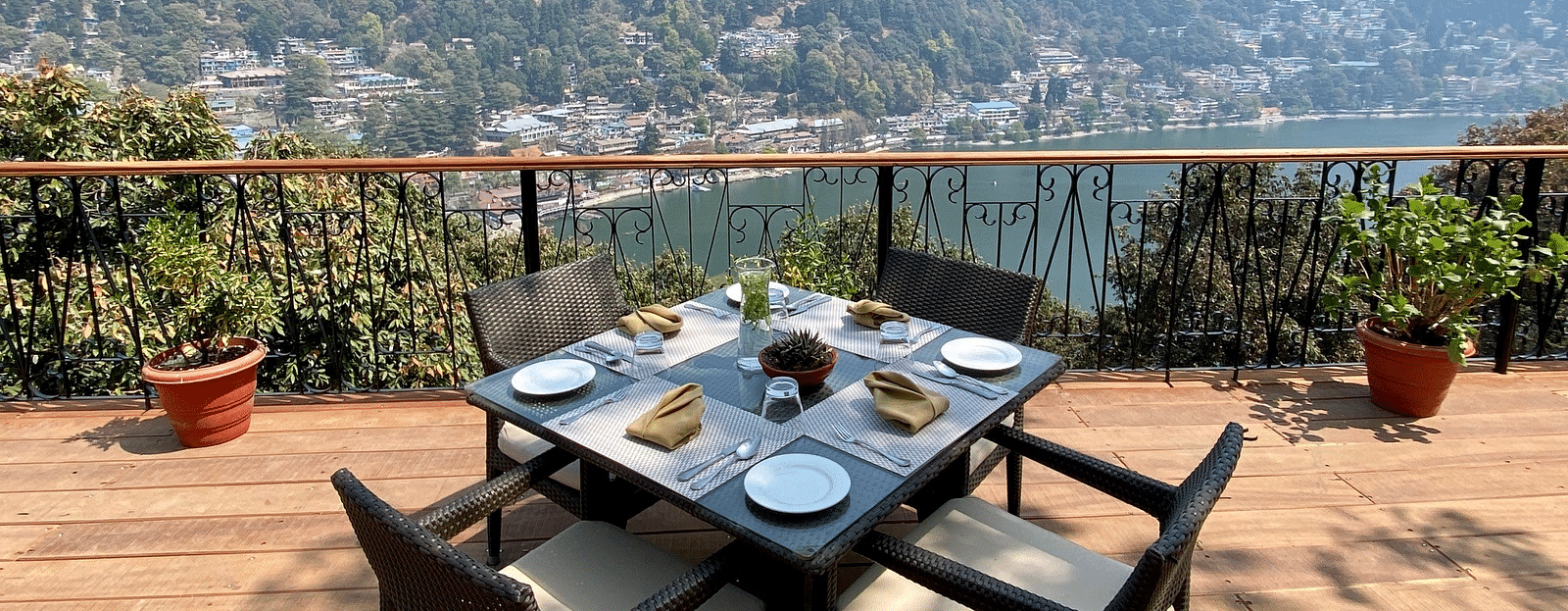 Image showing few chairs placed on the  sides of a square table, pot on either side of the frame, beautifully framed by the backdrop of the mountain ranges.