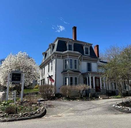 Hartstone Inn on a sunny day, showcasing its grand Victorian architecture, American flag on top, and blooming trees lining the driveway.