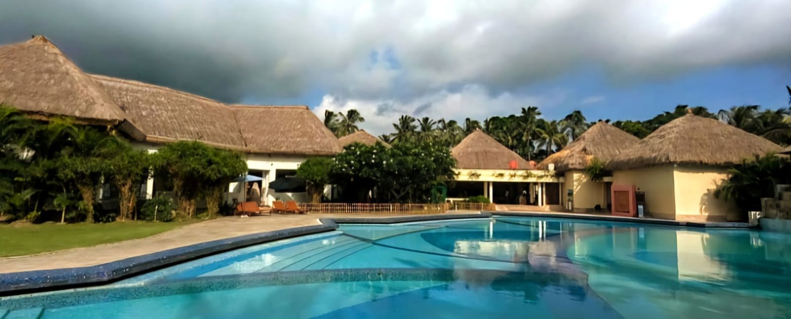 A wide view shot of Vedic Village Spa Resort featuring a swimming pool lined by buildings with thatched roofs under a partly cloudy blue sky.