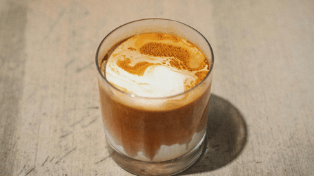 A glass of South Indian filter coffee topped with froth, served on a neutral surface in soft lighting.