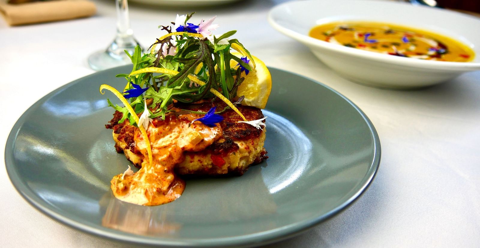 A detailed view of a fine-dining dish at Shaver Lake Village Hotel with fresh ingredients and a side of soup in the background.