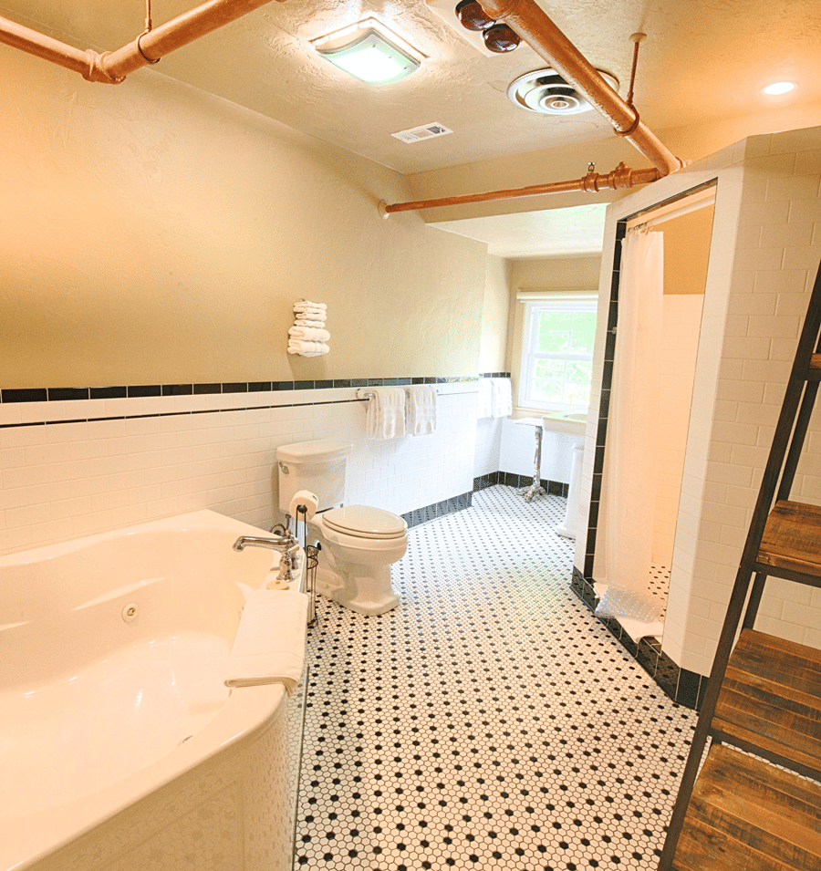 A modern bathroom with a white bathtub, toilet, tiled floor, exposed copper pipes, and a ladder