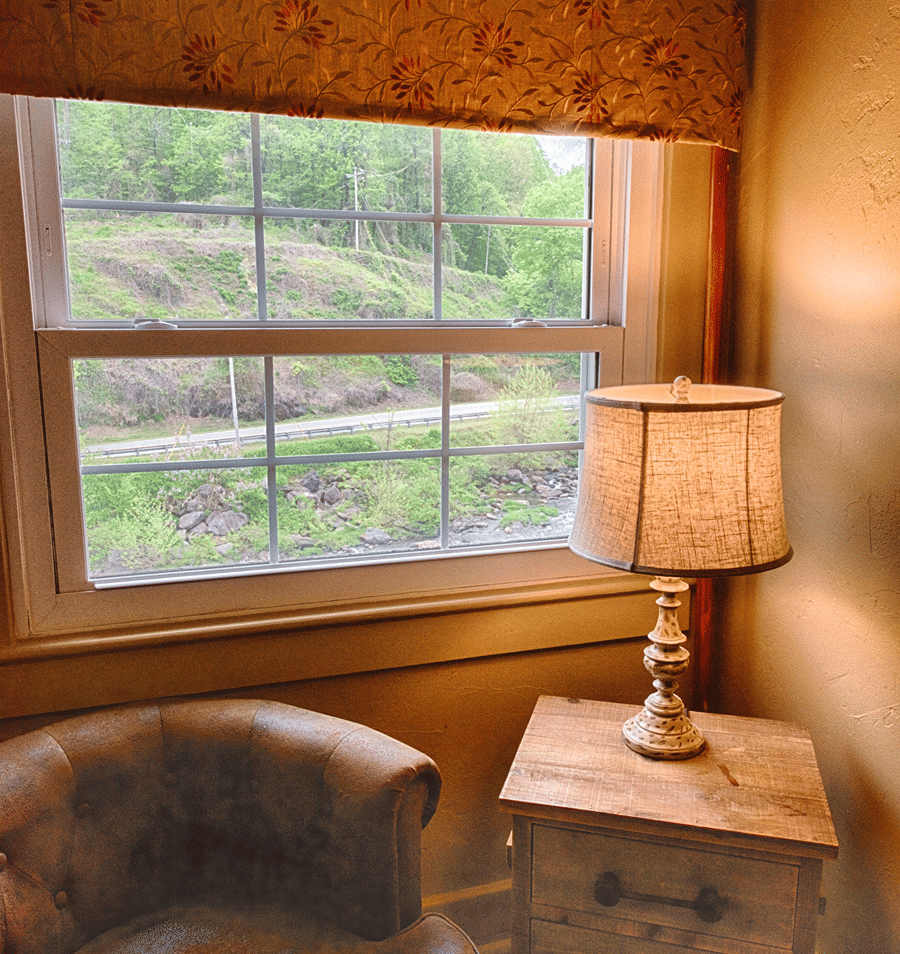 A cozy guest room with a large window showing a green outside view, a leather armchair, and a wooden nightstand with a table lamp.