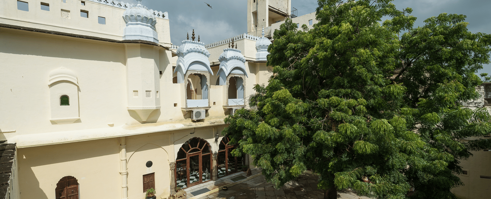 A courtyard at Fort Barli features a large tree, white buildings with decorative windows, and a distant tower against a cloudy sky.